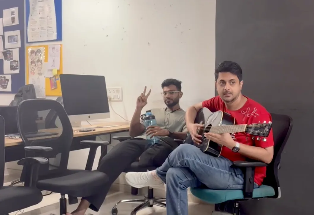 Playing an acoustic guitar in the studio while a colleague flashes a peace sign